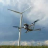 Drone flying near a large wind turbine in a field under a cloudy sky