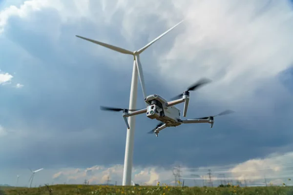 Drone flying near a large wind turbine in a field under a cloudy sky