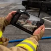 Firefighter holding a drone controller with a live thermal imaging feed on the screen during an outdoor operation